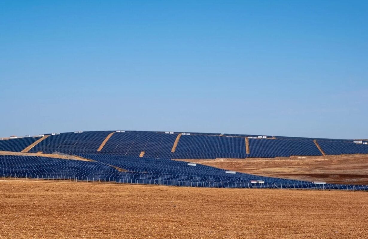 A large-scale solar panel farm under a clear sky in Turkey, with wind turbines visible in the background.