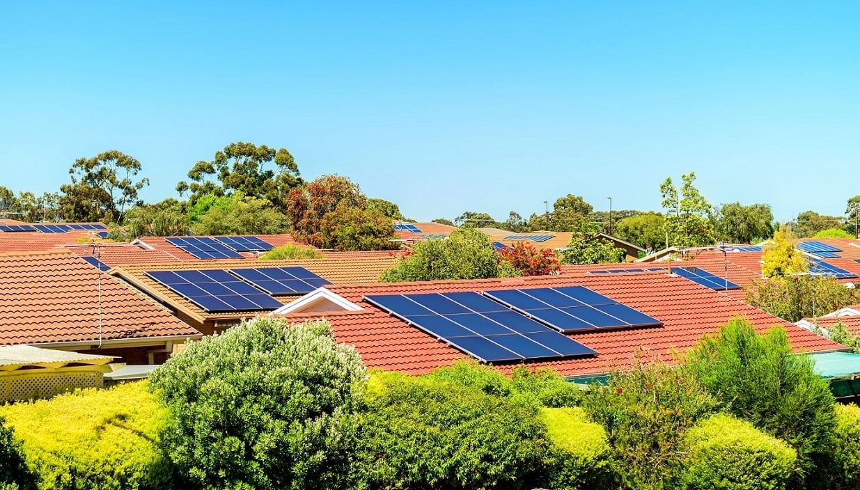 Aerial view of a large commercial warehouse with an empty roof next to residential homes with solar panels.