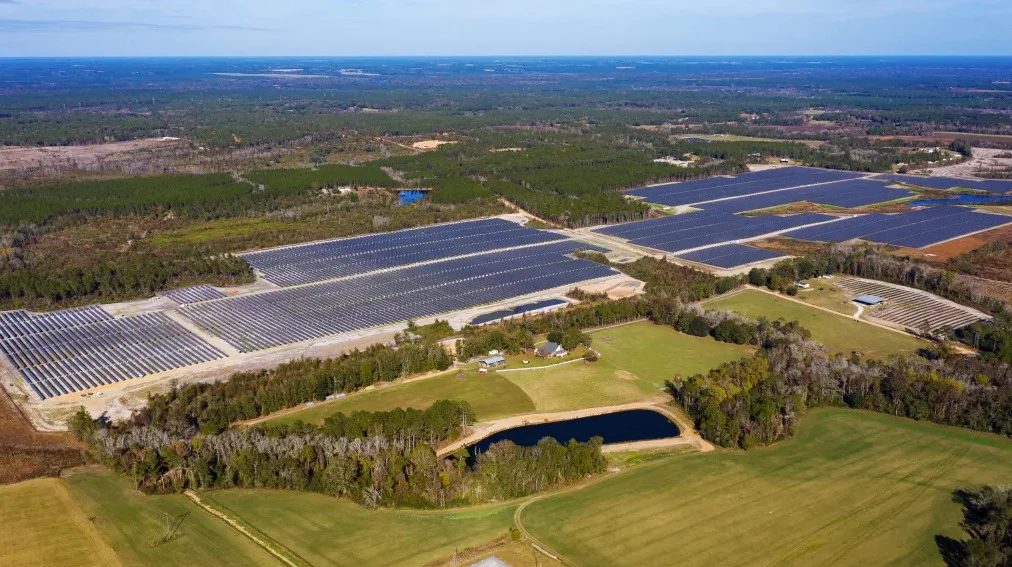 Aerial view of a massive solar array integrated with rows of white battery storage containers.