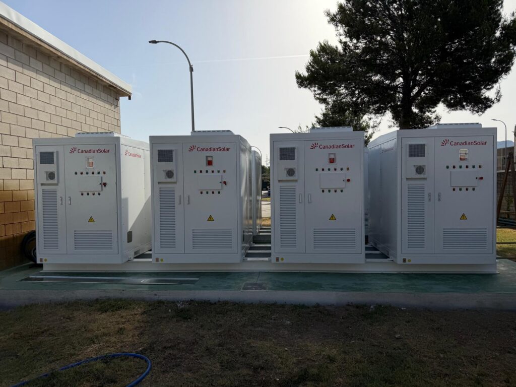 A large industrial rooftop solar installation with shipping container-sized battery storage units nearby.
