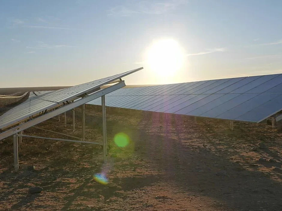 A massive utility-scale solar farm under a bright sun with high-voltage transmission lines in the background.