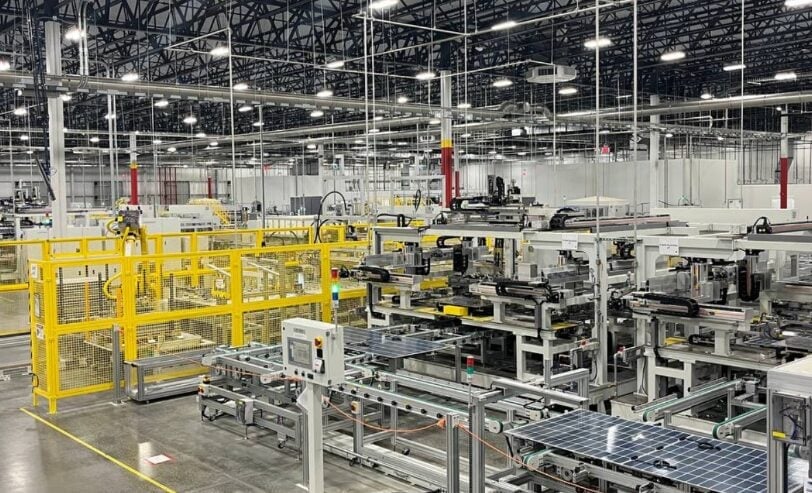 A large-scale solar panel manufacturing facility with workers and robotic assembly lines under bright lights.