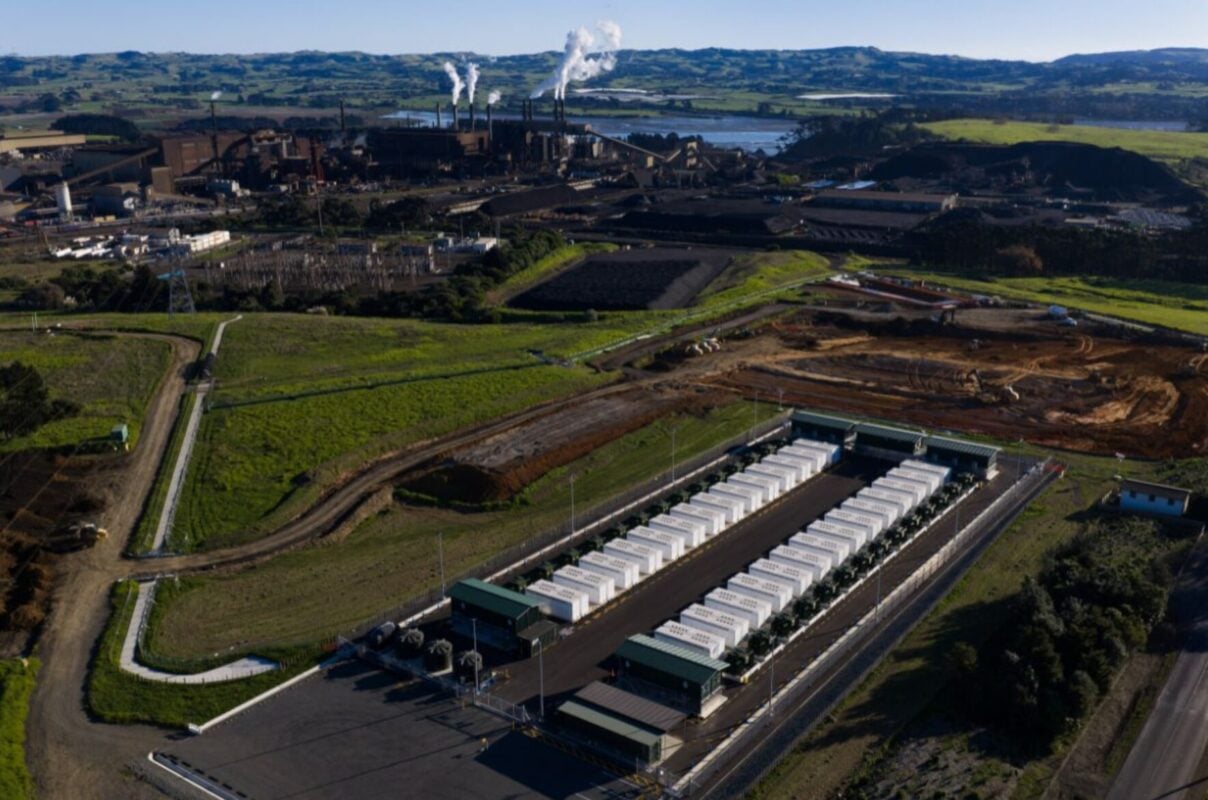 Large scale grid battery containers at an industrial site with steel mill infrastructure in background
