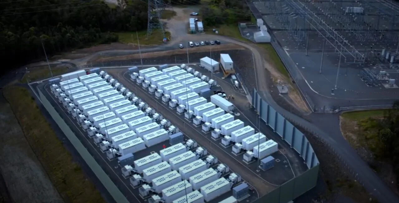 Industrial battery storage containers situated in a sun-drenched, arid landscape with solar panels in the background.